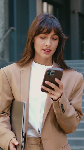 Close- up of woman with long hair holding smartphone in hands, looking at the camera with smiling against the background of modern building outdoors