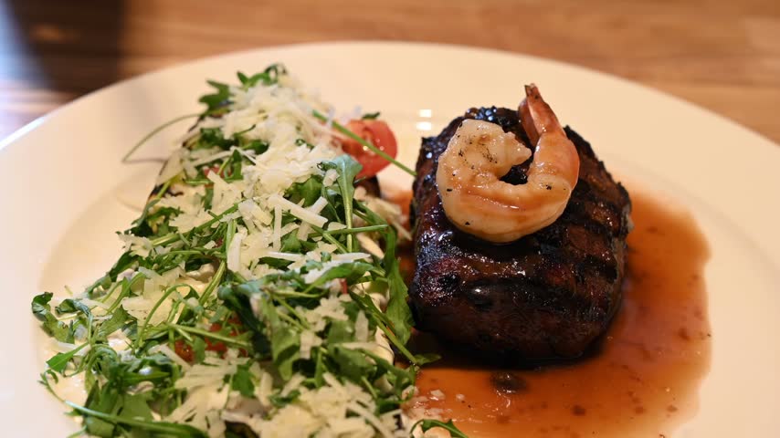Close up shot shows hand holding knife and fork slicing piece of juicy seasoned grilled beef steak garnished with shrimp served with fresh arugula salad on ceramic plate.