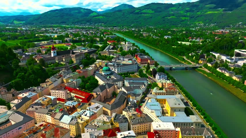 Aerial view of Salzburg city in Austria capturing beautiful historic architecture along Salzach river banks under bright summer sky surrounded by lush green hills scenic landscape drone shot.