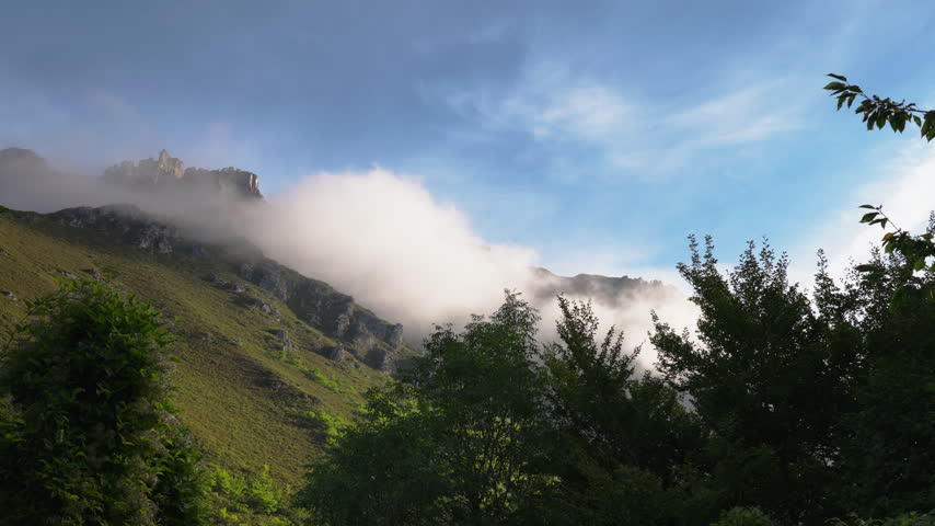 Stunning mountain landscape featuring low-lying clouds drifting swiftly across a green, rocky hillside, with lush trees in the foreground and a vibrant blue sky overhead