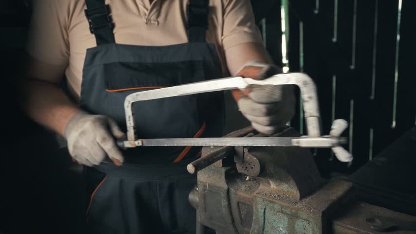 Skilled technician in work apron continues cutting metal pieces with a hacksaw, showcasing focused hand movements at a workbench surrounded by tools and materials in a dimly lit workshop setting