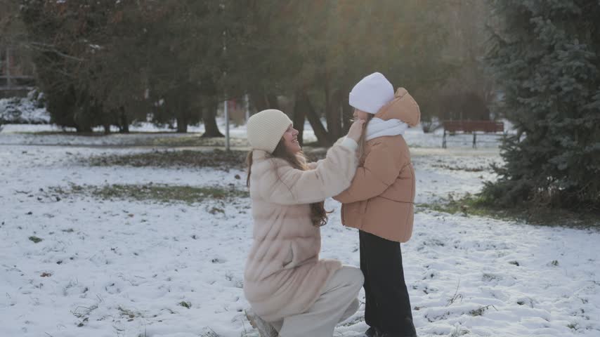 Young happy mother taking care of her daughter, zipping up her coat and talking to her during a walk in a beautiful snowy park