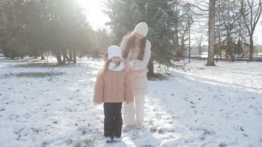 Two smiling sisters spending quality time together in a sunlit, snow-covered park. The older girl gently adjusts her younger sibling