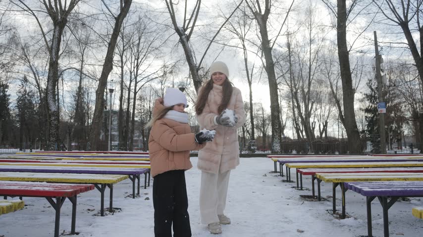 Two joyful sisters having fun together in a snowy park. Young girl and her older sister laughing, throwing snow, and playing together