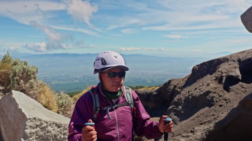 Middle aged man with helmet and poles hiking down a rocky mountain path on a sunny day