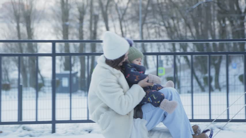 Happy young mother holding her infant son on her lap while gently swinging in a snowy park. Loving family enjoying a beautiful winter day