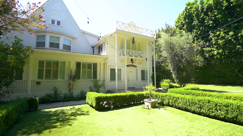 The camera moves toward a white two story house surrounded by trimmed hedges and a green lawn.