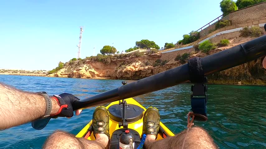 POV of person paddling yellow fishing kayak near rocky cliff coastline with promenade