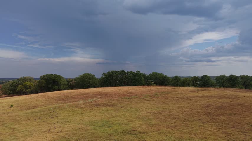 Rural Oklahoma farmland and hay fields revealing a distant rain cloud storm over treetops in an aerial rising forward shot.