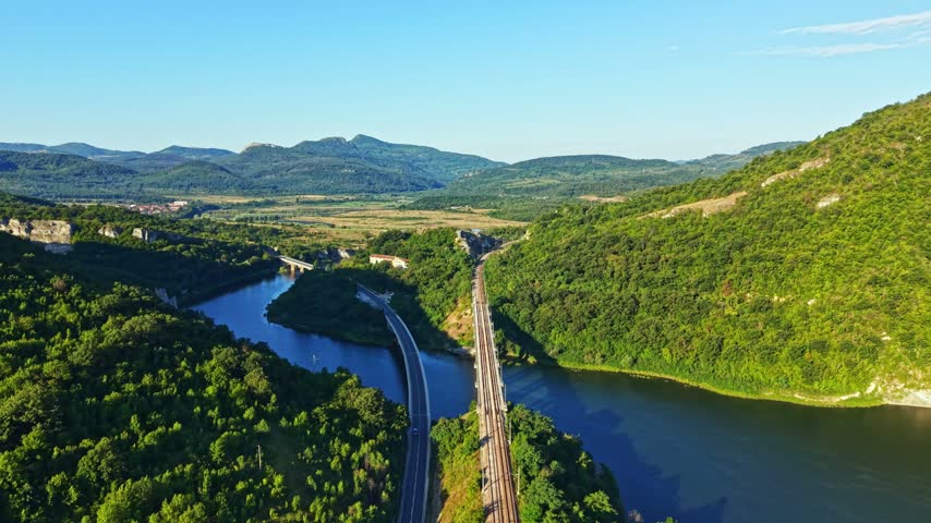 A landscape shows hills covered with trees and a river flowing beside railroad tracks. The sun is shining in the clear sky. This scene captures nature in a rural area.