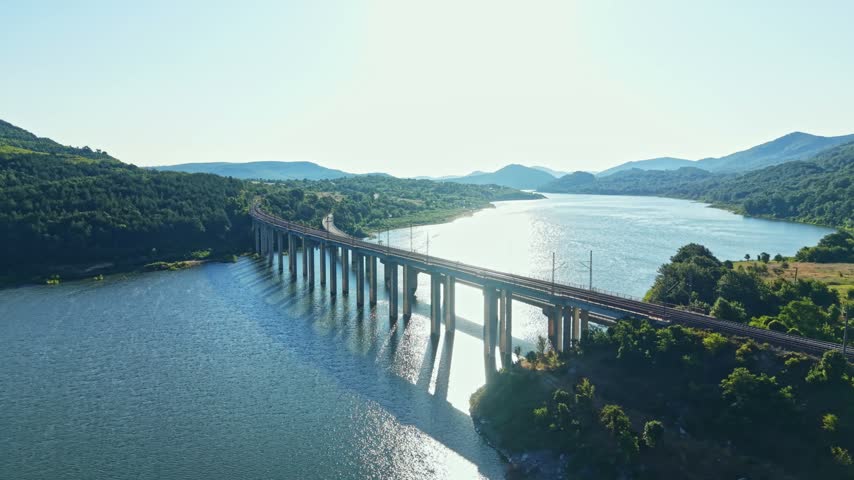 A bridge crosses a wide river, set against green hills in the background. The sun shines brightly above as train tracks run along the structure.