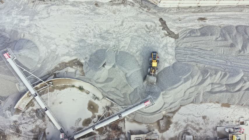 A yellow machine works in a granite quarry. The area is filled with large piles of stone gravel. Equipment is placed around the site. This occurs in a busy mining location.