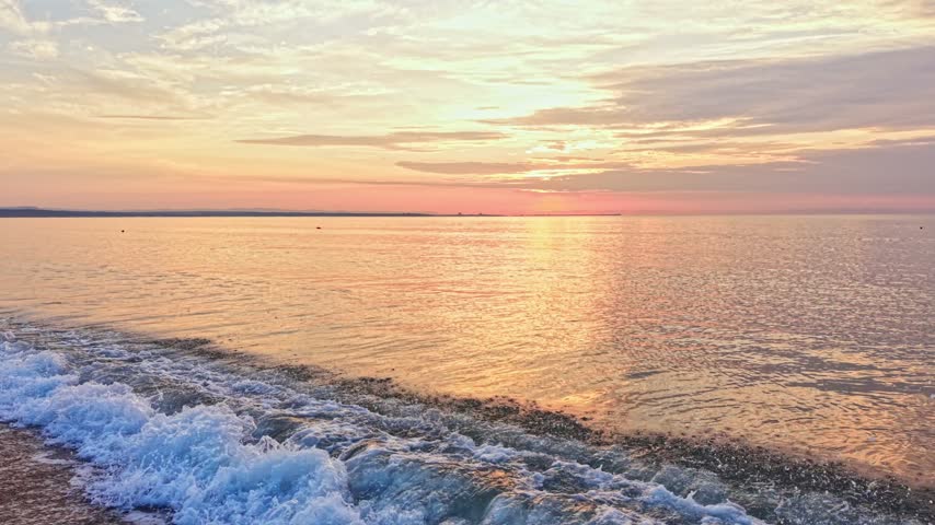 The sun sets over the ocean as waves gently roll onto the shore. The sky shows warm colors and clouds. This scene takes place during dusk by the water.
