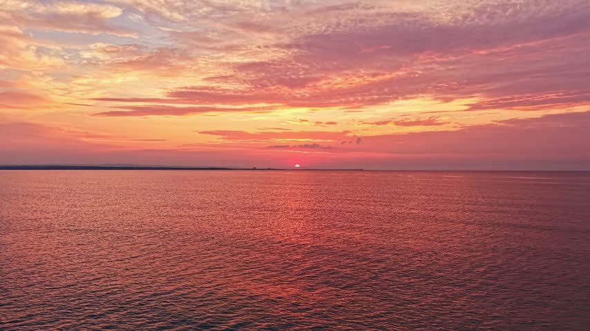 The sun sets on the horizon, casting shades of orange and pink across the sky. Calm water reflects the colorful sky. The scene is quiet and shows the beauty of nature in the evening.
