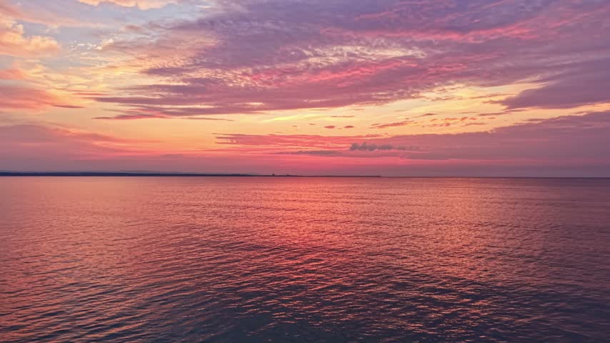 The sun sets over a body of water creating a mix of colors in the sky. Soft clouds spread out as the evening light reflects on the calm water nearby.