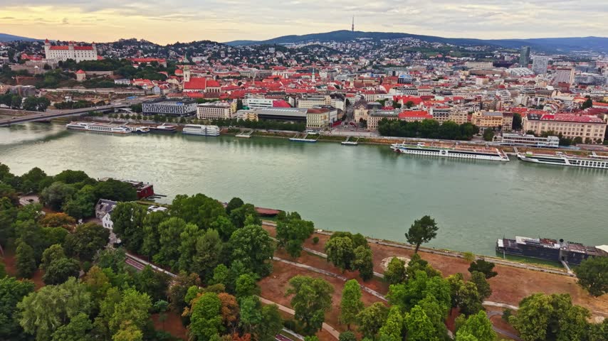 View of a city by the river with many buildings and trees during the late afternoon. The sky is cloudy, and there are boats on the water.