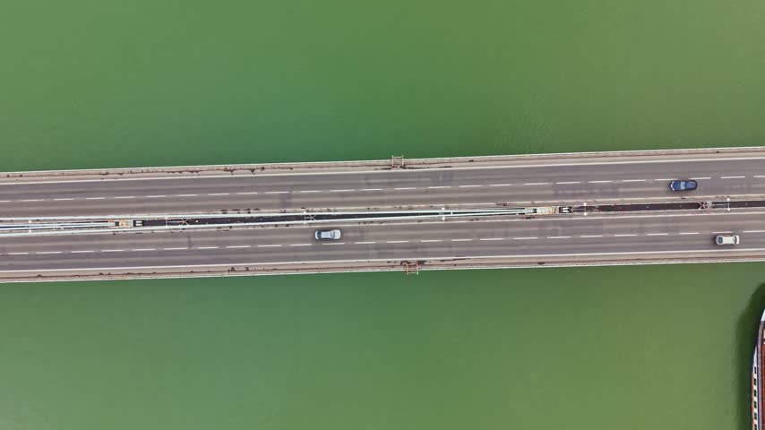 Cars move along a wide bridge that connects two land masses while boats float nearby in the water. The scene shows the flow of traffic and vessels in the area.