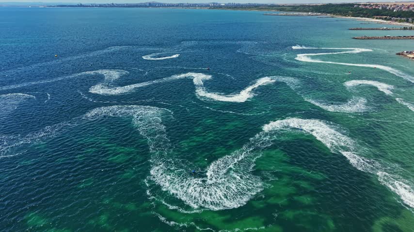 Riders on jet skis race across the water, creating waves and patterns. The scene takes place near the beach in a coastal city under clear skies and bright sunlight.