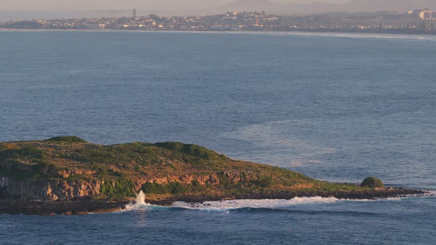 Footage of a small island with green vegetation, surrounded by blue ocean water as waves crash on its rocky shores, and a distant coastline with buildings under a hazy sky.
