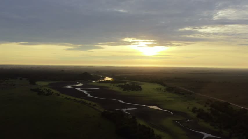 Sunlit marsh network glowing under a low rising sun over the dark silhouetted Orinoquia savanna, aerial pullback drawing away from the water