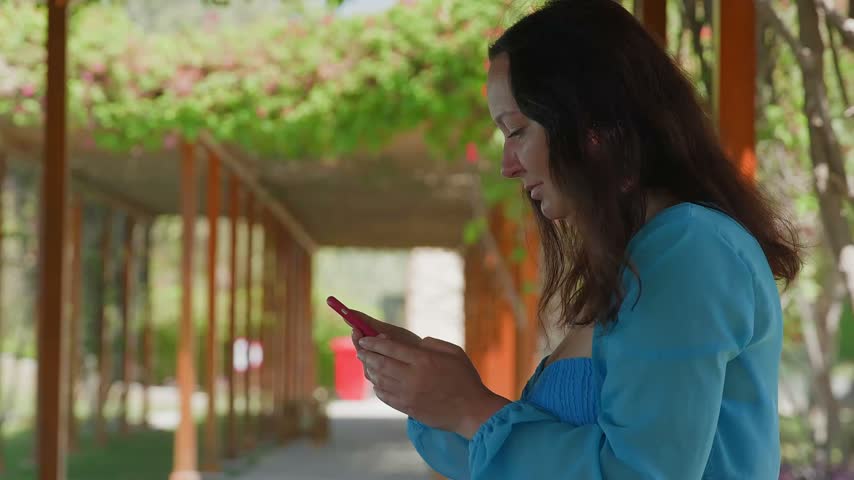 Woman in blue shirt holds smartphone while standing under blooming flowering vines in covered garden walkway with wooden structure, natural daylight filtering through foliage.