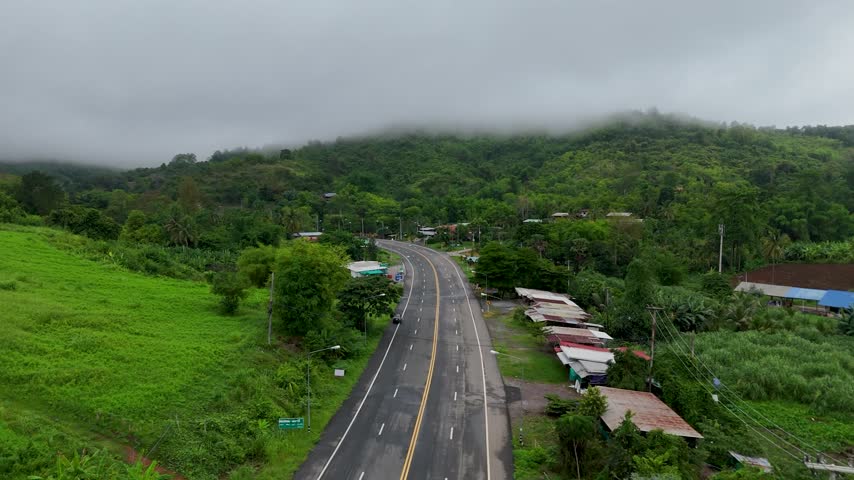 Aerial drone footage capturing rural highway curves passing through tropical greenery and misty mountains. Scenic landscape showcases moody weather patterns with low clouds hanging over hillside scenery.