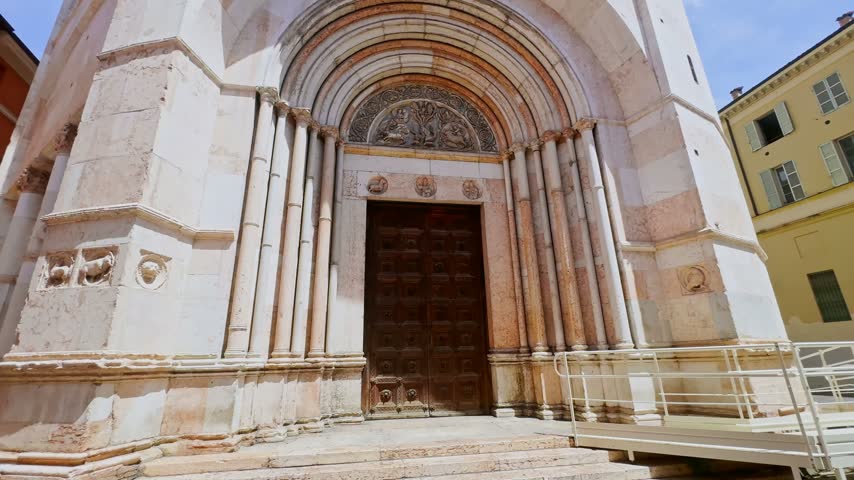 Front view of the Baptistery of Parma entrance, showing its carved Romanesque marble portal, wooden doors, sculpted tympanum and accessibility ramp in Parma, Italy.