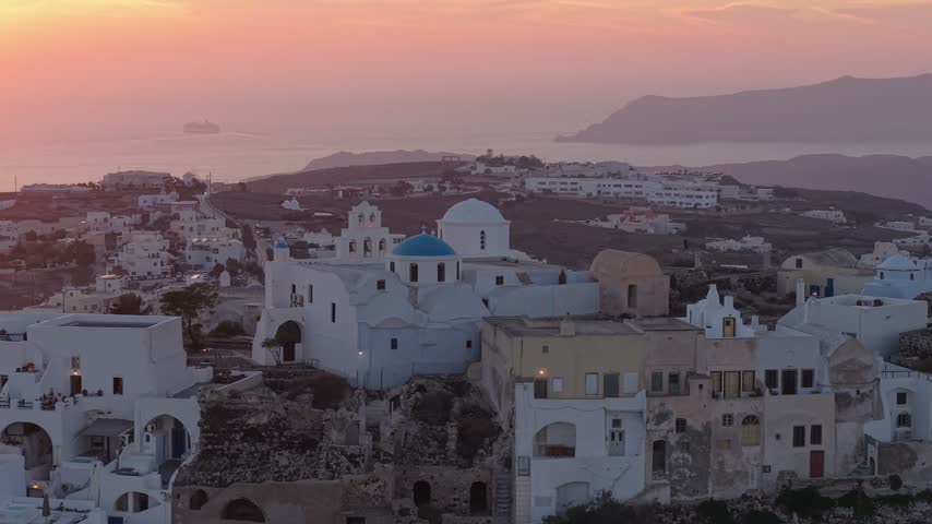 Breathtaking aerial view over traditional Santorini village with blue domes and white buildings during golden sunset hours. Peaceful Greek island scenery with calm sea and distant silhouettes at dusk.