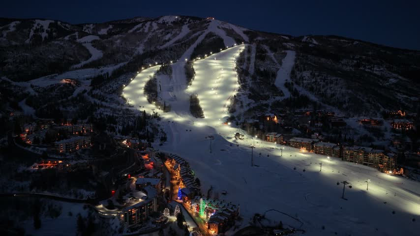 Night aerial of a snowy mountain resort resembling Breckenridge, Colorado, with illuminated ski slopes, active night skiing, and a glowing town of hotels and homes at the base after dark.