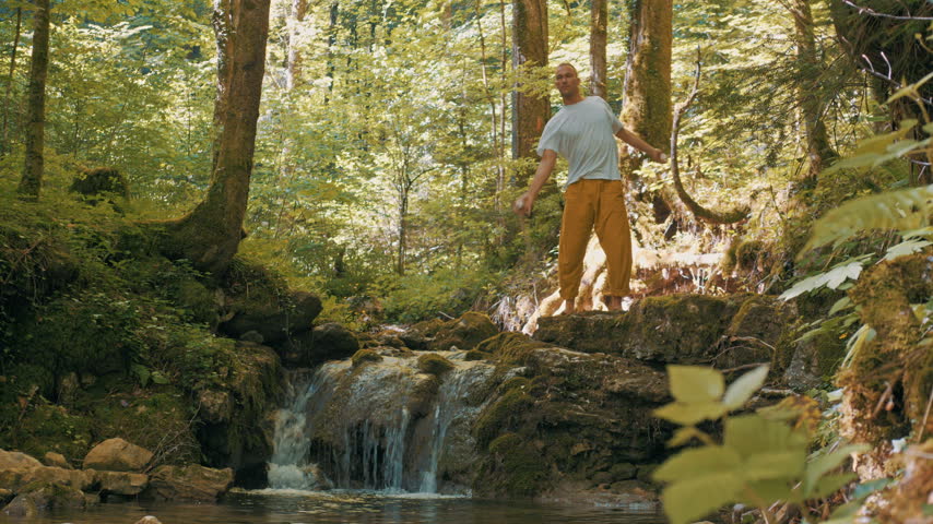 A man performs a balance exercise on a rocky outcrop beside a cascading waterfall in a lush forest.