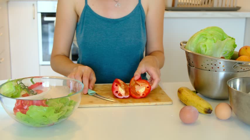 Unrecognizable woman slices sweet red bell pepper into small pieces on wooden board. Glass bowl with mixed green lettuce and red tomatoes stands on white table during healthy cooking.