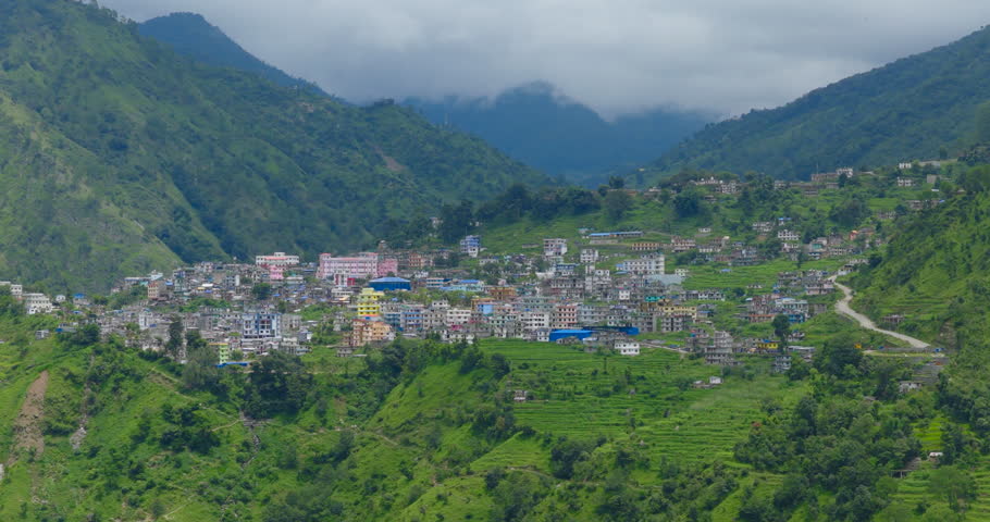 Done shot of Martadi, Bajura district, Nepal, of the suburban district headquarters settlement spread across vibrant green hillsides and terraced fields beneath cloud-draped mountains peaks