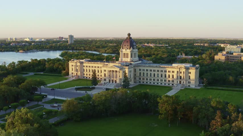 Cinematic drone shot approaching historic limestone parliament house. Lush green park landscape surrounding government landmark. Golden hour lighting over scenic urban lake and city horizon.