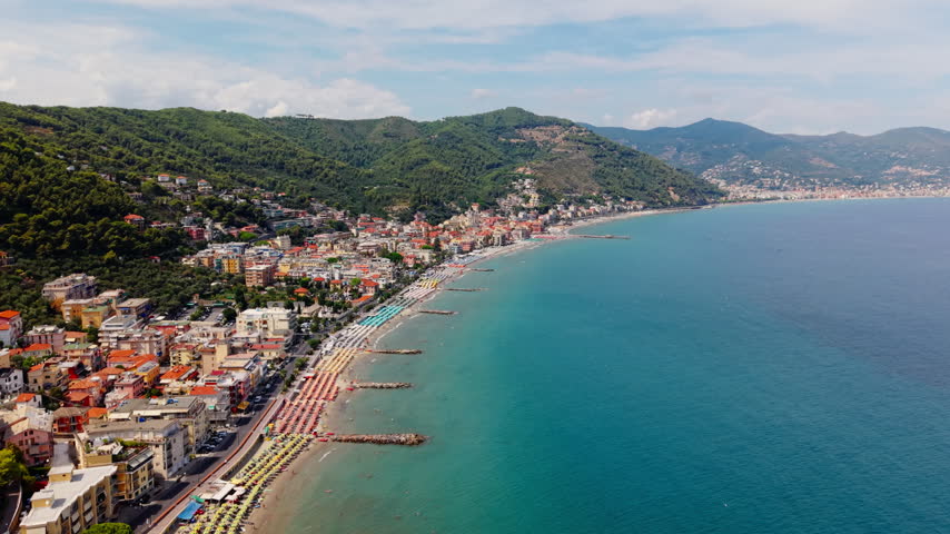 Aerial drone shot retreating along the Laigueglia coastline, revealing beach umbrellas, sun loungers, seaside buildings, breakwaters and green hills behind the town.