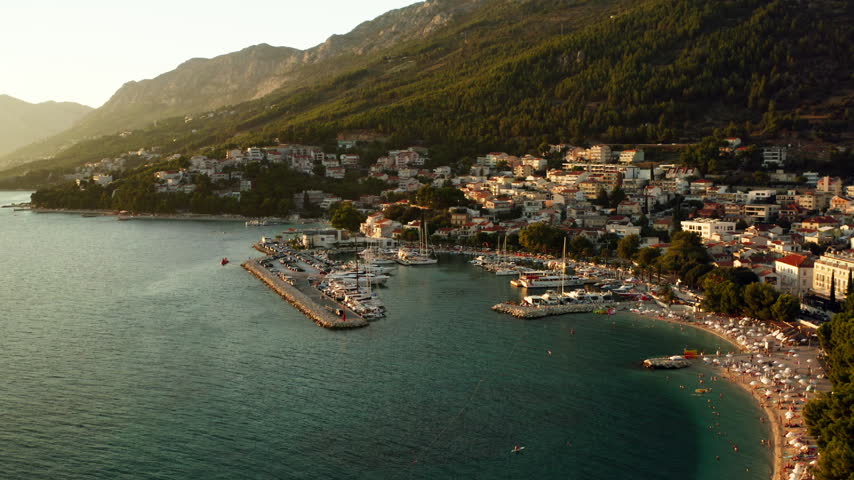 The marina harbor of Baska Voda in Croatia shelters moored boats along the stone pier. The Adriatic coastal town includes a pebble beach, residential houses, and forested mountain slopes.