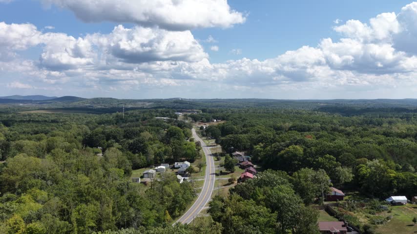 A two lane highway threads through wooded countryside outside Lynchburg, Virginia, scattered houses along the verge and rolling forest running back to the horizon. Aerial forward wide shot
