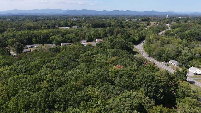 A flight along the wooded outskirts of Lynchburg, Virginia passes houses, small commercial buildings, open pasture and a farm pond, the Blue Ridge low on the horizon. Aerial flyover shot