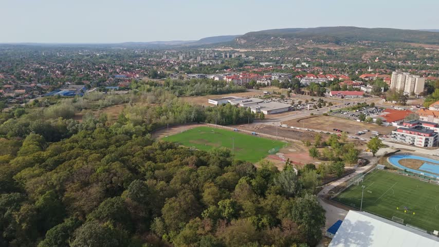 Tatabánya sports complex with football pitch and athletics track beside residential areas and urban greenery, with forested hills and roads visible in summer, filmed by drone.
