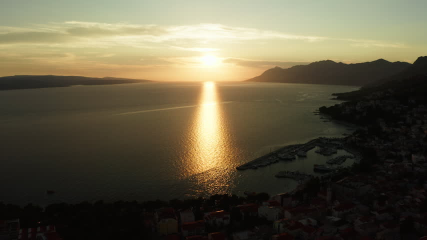 The sun sets over the calm Adriatic Sea in Baska Voda, Croatia. Bright solar reflection shines on the water near the coastal resort harbor. Peaceful summer evening brings tranquil holiday mood.