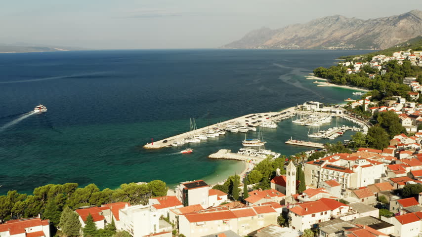 Baska Voda coastal resort town features a sheltered yacht marina harbor in Croatia. Boats moor along the Adriatic Sea waterfront beside dense red roof houses on the Makarska Riviera coast.