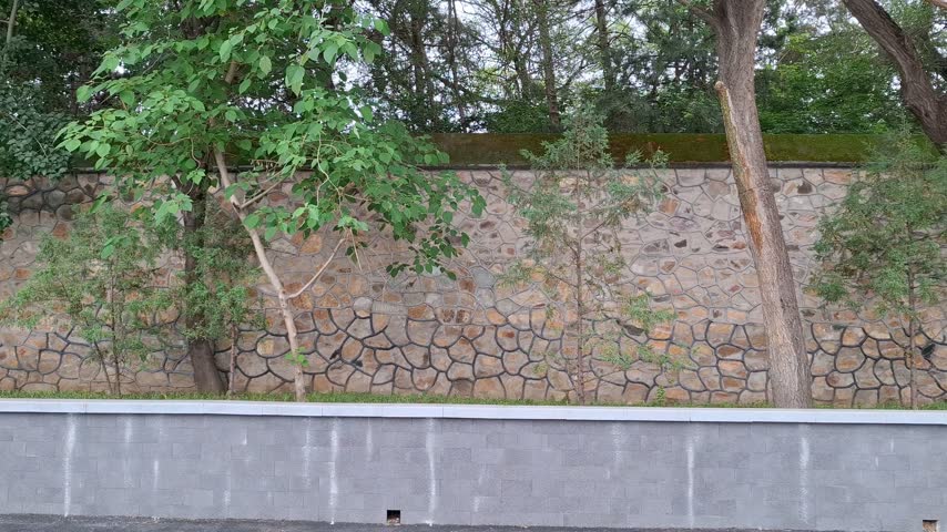 Ancient traditional stone wall with moss growing on top behind fresh green trees in a park, historic oriental garden architecture and travel concept.