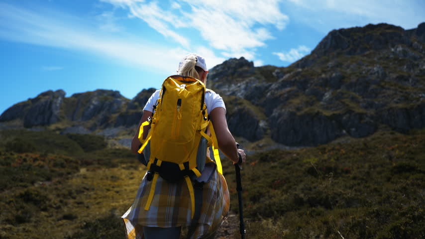 Blonde woman hiker with a yellow backpack and trekking poles walking away from the camera on a mountain path, enjoying an active outdoor adventure in a beautiful natural landscape