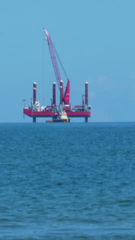 A red jackup rig or offshore construction vessel with a large crane stands in the blue waters of the Baltic Sea near Osetnik, Poland, representing energy and industrial infrastructure.