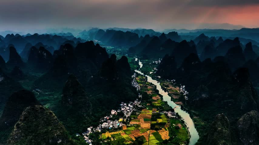 An elevated high-angle aerial view captures the scenic Li River winding through the dramatic limestone karst mountain peaks of Guilin and Yangshuo in Guangxi, China. The expansive composition surveys dark conical rock formations flanking a deep water channel under twilight and evening lighting conditions. This regional landscape asset suits documentary features, Asian tourism promotions, geographical establishing sequences, and atmospheric environmental b-roll.