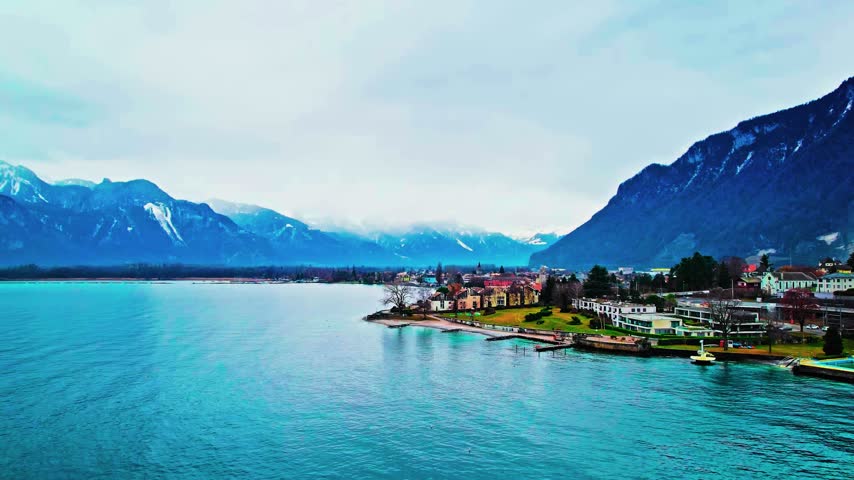An elevated high-angle aerial view captures a lakeside town along Lake Geneva in the Swiss Riviera region of Switzerland. The expansive composition surveys residential buildings, urban waterfront properties, a paved coastal shore, and dramatic alpine mountain ranges under overcast daylight conditions. This travel and regional landscape asset suits documentary features, European tourism promotions, geographical establishing sequences, and cinematic aerial b-roll requiring authentic Swiss lake geography.