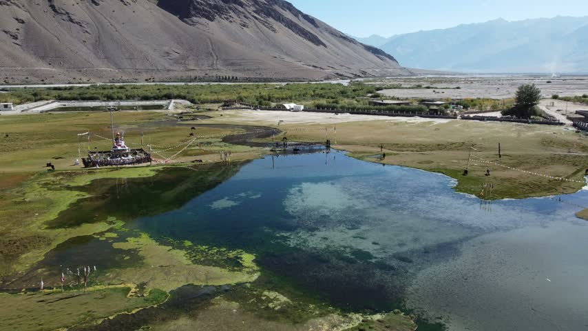 The video showcases a serene aerial view of Sani Lake in Padum, India. The tranquil lake is surrounded by lush greenery and dotted with prayer flags fluttering in the breeze.