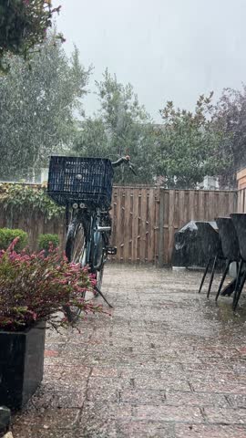 Heavy rain falls in a backyard where a bicycle with a front basket stands near a barbecue table.