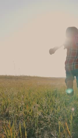 A boy walks through tall grass in an open field. The boy enjoys the peaceful scenery, exploring while the sun sets in the background.