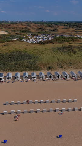 Vertical aerial drone video shot looking down over the wide sandy resort at Katwijk aan Zee, South Holland, Netherlands. The camera movement traces a long row of uniform white houses and beach huts lined up along the foot of grassy coastal sand dunes. On the golden sand, rows of beach chairs, windbreaks, and sunbathers enjoy a warm sunny summer afternoon. Beyond the primary coastal dunes, a large seaside campsite with camper vans and caravans sits in the grassy dune landscape, leading toward distant city skyline views under a clear blue sky. High-resolution stock media highlighting Dutch coastal beach tourism, beach house rentals, dune conservation areas, and North Sea seaside recreation. Ideal for vertical social media, mobile video production, travel stories, commercial advertising, and broadcast media.