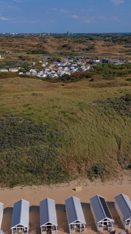 Vertical aerial drone video shot looking over rolling coastal sand dunes and seaside resorts at Katwijk aan Zee, South Holland, Netherlands. The camera movement rises above a row of white wooden beach houses and beach huts situated on the edge of the sandy beach shoreline. Above the dunes, covered in marram grass and coastal vegetation, a large seaside campsite with camper vans, caravans, and tents sits nestled in the green landscape. In the background, expansive coastal dune reserves lead toward the distant city skyline under a clear blue sky on a bright summer afternoon. High-resolution stock media highlighting Dutch beach tourism, dune conservation landscapes, camping holidays, and North Sea coastal nature. Ideal for vertical social media reels, mobile video production, travel stories, commercial advertising, and broadcast media projects.